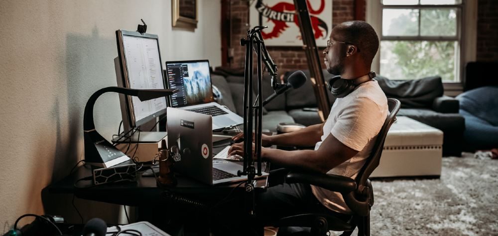 Third Culture Africa 6 Essential Pieces Of Podcast Equipment You Need For Success a man wearing a cream t-shirt sitting on a brown office chair in front of a desk with speakers attached and laptop and mixer on the table with headphones on his neck