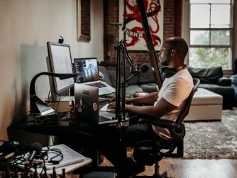 Third Culture Africa 6 Essential Pieces Of Podcast Equipment You Need For Success a man wearing a cream t-shirt sitting on a brown office chair in front of a desk with speakers attached and laptop and mixer on the table with headphones on his neck