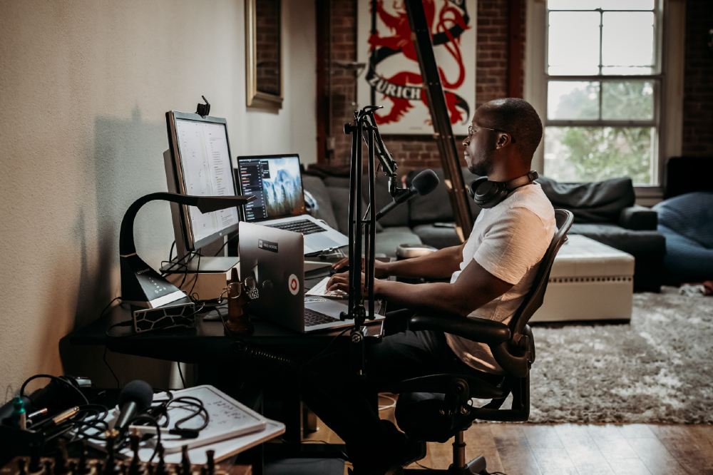 Third Culture Africa 6 Essential Pieces Of Podcast Equipment You Need For Success a man wearing a cream t-shirt sitting on a brown office chair in front of a desk with speakers attached and laptop and mixer on the table with headphones on his neck
