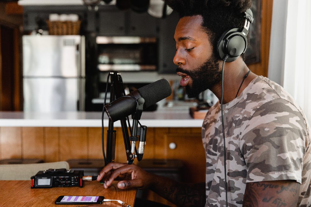 Third Culture Africa How Do I Podcast a man wearing a beige t-shirt with tattoos over his arms has earphone on and podcasting on a microphone attached to a brow table with a cellphone on top of the table