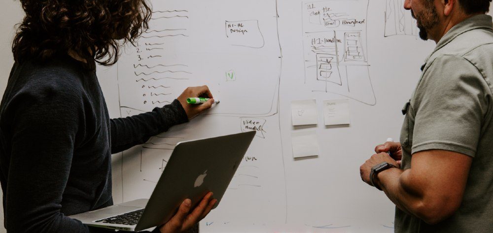 Third Culture Africa Leadership Traits For Success a woman holding laptop in her right and writing on a white board with her left hand using green marker and a man paying attention to her writing.