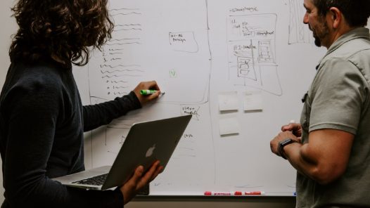 Third Culture Africa Leadership Traits For Success a woman holding laptop in her right and writing on a white board with her left hand using green marker and a man paying attention to her writing.
