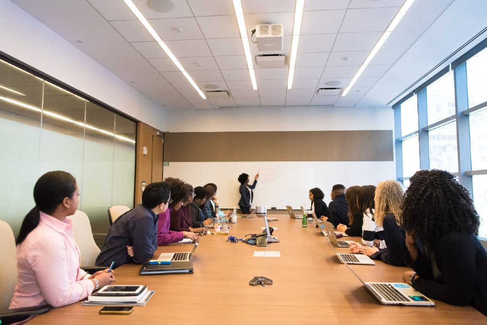 Third Culture Africa Transformational Leadership a group of people in boardroom with laptops and note books on a brown table all facing one direction to a woman writing on a white board
